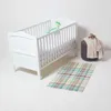 A white baby crib with a green toy dinosaur inside, next to a gray ottoman with blankets and a plaid rug on a white floor. Produktbild-Vorschau