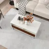 A white coffee table with two drawers and open shelves is placed on a light gray rug in a living room setting. A sofa with a black and white throw blanket is visible in the background. Produktbild-Vorschau