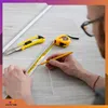 A person's hands are using a tape measure and pencil to mark a white strip on a tiled floor, with a utility knife and tape measure nearby. Produktbild-Vorschau