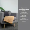 A beige fleece blanket draped over a gray armchair, with a dark gray cushion and a green plant leaf visible. Náhľad obrázku produktu