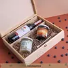 A wooden gift box containing a bottle of red wine, a jar of pasta sauce, a jar of coconut oil, and a honey dipper, surrounded by blueberries. Produktbild-Vorschau