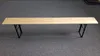 A wooden bench with black metal legs is shown on a gray carpeted floor. Náhled obrázku produktu