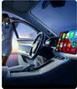 A woman is leaning against the open door of a car, holding a smartphone. The car's interior is visible, featuring a large touchscreen display showing various app icons. Produktbild-Vorschau
