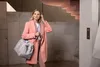 A woman stands in a modern building lobby, holding a duffel bag and adjusting her hair. Produktbild-Vorschau