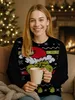 A young woman wearing a Grinch-themed Christmas sweater holds a mug in a cozy, festive setting with a Christmas tree and fireplace. Produktbild-Vorschau