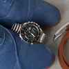 A close-up shot of a men's watch and blue suede shoes, with a brown leather belt and buckle visible on the right. Produktbild-Vorschau
