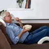 An elderly man is relaxing on a brown sofa, eating popcorn and watching TV. He is using a white armrest pillow. Anteprima dell’immagine prodotto
