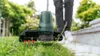 A person is using a green string trimmer to cut grass next to a stone pathway. Produktbild-Vorschau