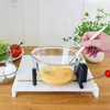 A person is using a manual egg beater to whisk eggs in a glass bowl, which is placed on a white cutting board with suction cups. Other ingredients like lettuce and tomato are visible in the background. Produktbild-Vorschau