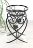 A black metal plant stand with decorative scrollwork and leaf motifs, shown in a studio setting with a blurred green plant in the background. Náhled obrázku produktu