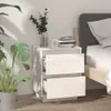 A white wooden nightstand with two drawers next to a bed, with a plant in a pot and a vase of greenery on top. Náhled obrázku produktu