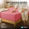 A person sitting on a pink bed sheet on a wooden bed frame, with a bedside table and lamp in the background. Produktbild-Vorschau