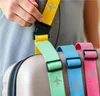 A person's hands are shown attaching a yellow luggage strap with an airplane icon to a suitcase. Three other colorful luggage straps (green, blue, and pink) are also visible, attached to the same suitcase. Podgląd zdjęcia produktu