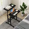 A black adjustable laptop table with a laptop, books, and a coffee cup on it, placed next to a bed and a large potted plant. Podgląd zdjęcia produktu