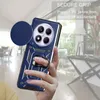 A hand holding a blue phone case with a camera cover and ring holder, with a blurred background of an indoor setting. Produktbild-Vorschau
