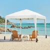 A white pop-up canopy tent is set up on a sandy beach next to the ocean, with two wooden chairs and a small table inside. Produktbild-Vorschau