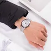 A person's wrist wearing a silver watch with a white face, with a black jacket sleeve visible. Produktbild-Vorschau