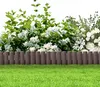 A garden border made of brown plastic stakes with a wood-like texture, separating a lush green lawn from a dense bush with white flowers. Náhled obrázku produktu