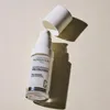 A white bottle of serum with its cap off, casting a shadow on a light-colored surface. Produktbild-Vorschau