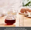 A glass teapot with red tea and another glass teapot with clear liquid are on a wooden table, with a cake under a glass dome in the background. Produktbild-Vorschau