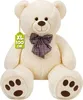 A large, cream-colored teddy bear with a plaid bow tie is shown against a white background. A green tag with text is attached to its left ear. Náhľad obrázku produktu