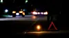 A warning triangle and a flashing warning light are placed on the side of a road at night, with blurred car headlights in the background. Produktbild-Vorschau
