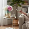 A person is using a smartphone next to a side table with a vase of flowers and a cup of coffee. Produktbild-Vorschau