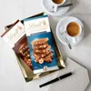 Two Lindt chocolate bars are displayed on a gold tray next to two cups of coffee and a pen. Produktbild-Vorschau