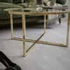 A close-up view of a round glass-top coffee table with a gold metal frame, placed on a light gray carpet in a living room setting. A green sofa is visible in the background. Náhľad obrázku produktu