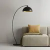 A black arc floor lamp is positioned next to a beige sofa with a yellow pillow and a green throw blanket. Produktbild-Vorschau