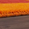 A close-up view of the edge of a textured rug with horizontal stripes of orange and red, resting on a dark wooden floor. Produktbild-Vorschau