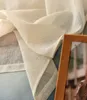 A close-up of a sheer, cream-colored curtain with a textured weave, draped over a light blue surface. A wooden picture frame is partially visible on the right side. Produktbild-Vorschau