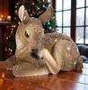 A decorative fawn figurine covered in artificial snow sits on a wooden table in front of a Christmas tree and a window. Produktbild-Vorschau