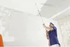 A person is using a spray applicator to apply material to a ceiling in an unfinished room. Produktbild-Vorschau