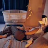 A person is lying on a bed next to a baby bassinet, with musical notes floating in the air. Anteprima dell’immagine prodotto