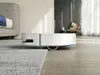 A white robot vacuum cleaner is shown on a floor next to a rug, with a sofa in the background. Náhled obrázku produktu