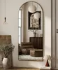 A full-length arched mirror reflects a stylish living room interior with a brown armchair and ottoman, a dark wood dresser, and decorative lighting. Anteprima dell’immagine prodotto