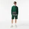 A man wearing a green Lacoste outfit and a black bag stands against a white background. Voir les images du produit