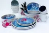 A collection of blue and white ceramic tableware, including bowls, plates, and mugs, arranged on a white surface with pink flowers and dark foliage. Produktbild-Vorschau