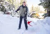 A man is shoveling snow in a snowy environment with trees and a cabin in the background. Náhled obrázku produktu