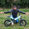 A man is kneeling next to a light blue electric bicycle in a grassy outdoor setting. Produktbild-Vorschau