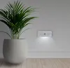 A white motion-activated LED night light is mounted on a white wall next to a potted plant. Produktbild-Vorschau