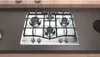 A stainless steel gas hob with four burners is installed on a dark countertop, with wooden cabinets visible in the background. Náhľad obrázku produktu