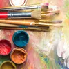 A collection of paintbrushes and small containers of paint are arranged on a colorful, textured surface that appears to be a painter's palette. Produktbild-Vorschau