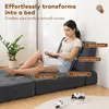 A woman sits on a modular sofa that is configured as a lounge chair, reading a book. The sofa is dark gray and appears to be made of corduroy. The background is a plain wall with a window and a plant. There are also game controllers on the floor. Produktbild-Vorschau
