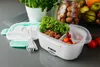 A white electric lunchbox filled with food, placed on a gray countertop next to its lid and a power cord. In the background, there are plants and a jar of nuts. Náhľad obrázku produktu