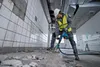 A construction worker is using a demolition hammer in a room with tiled walls and a debris-covered floor. Produktbild-Vorschau