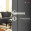 A door handle set is shown on a dark gray door, with a blurred living room in the background. Produktbild-Vorschau