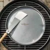 A round stainless steel grill plate with a spatula on top, placed on a grill grate. Podgląd zdjęcia produktu