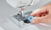 A hand is holding a blue bobbin near the needle of a sewing machine. Produktbild-Vorschau